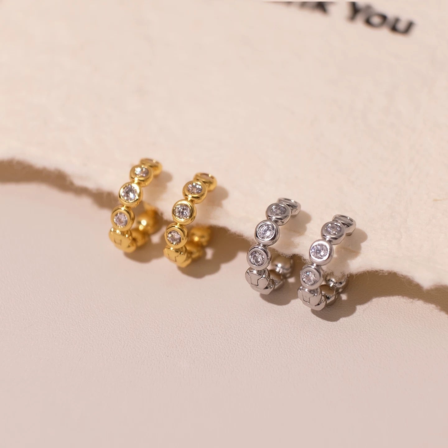 Gold and silver Elara bezel huggie earrings displayed together on a blush background