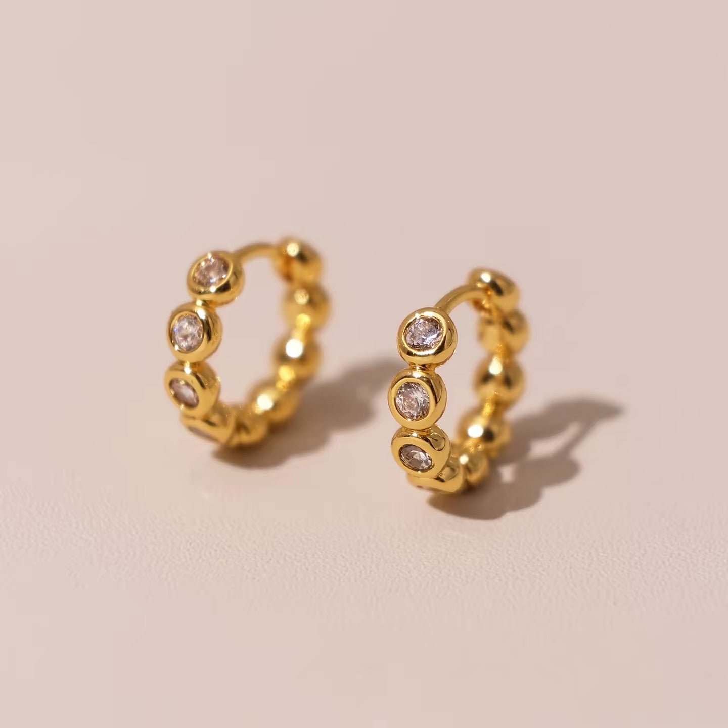 Gold Elara huggie earrings with sparkling bezel-set stones on a blush background