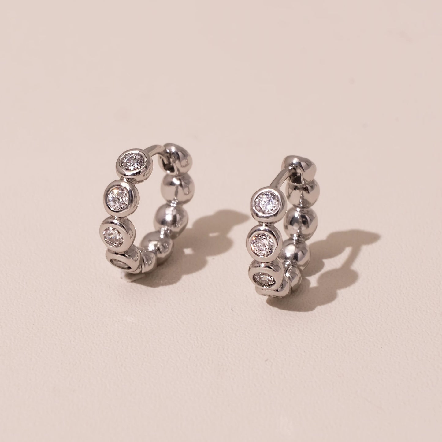 Pair of silver bezel-set huggie earrings photographed on a soft blush background
