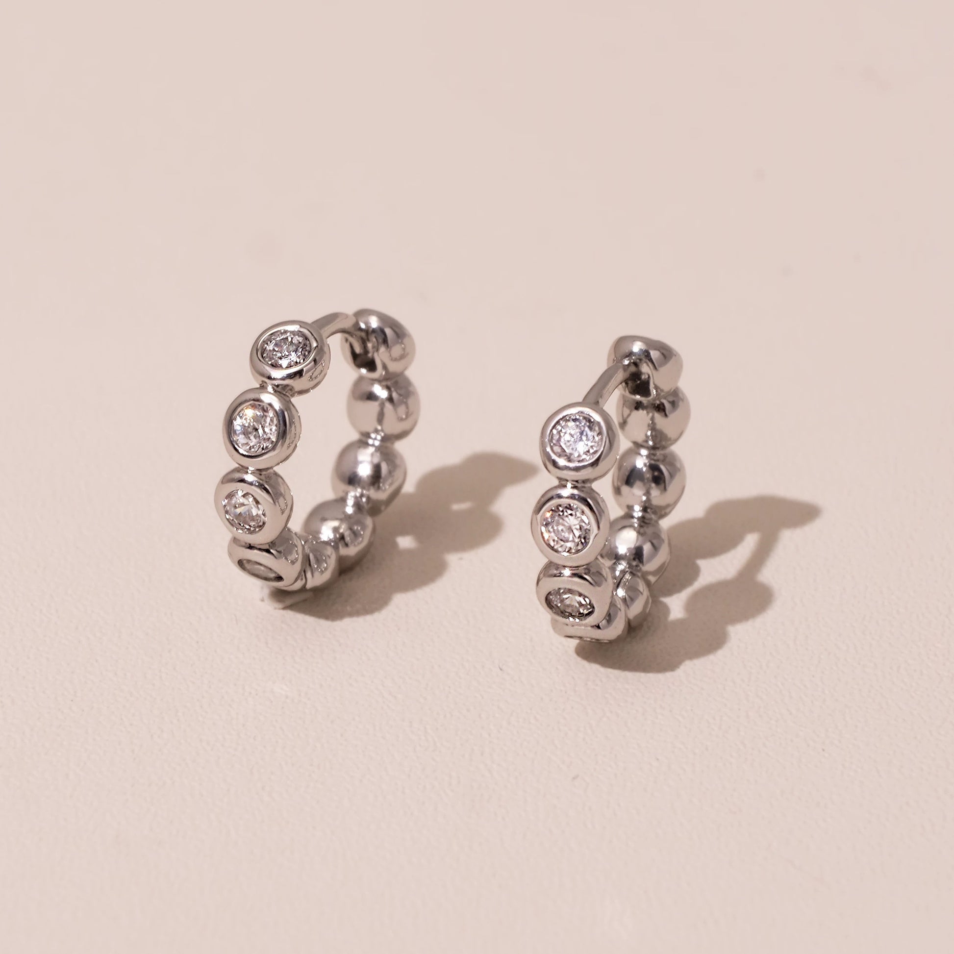 Pair of silver bezel-set huggie earrings photographed on a soft blush background