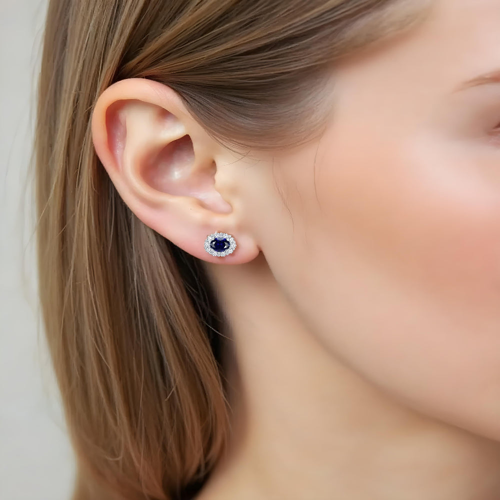 Model wearing Aeterna Jewelry blue sapphire halo stud earrings, close-up ear view showing deep blue gemstone surrounded by sparkling diamond halo